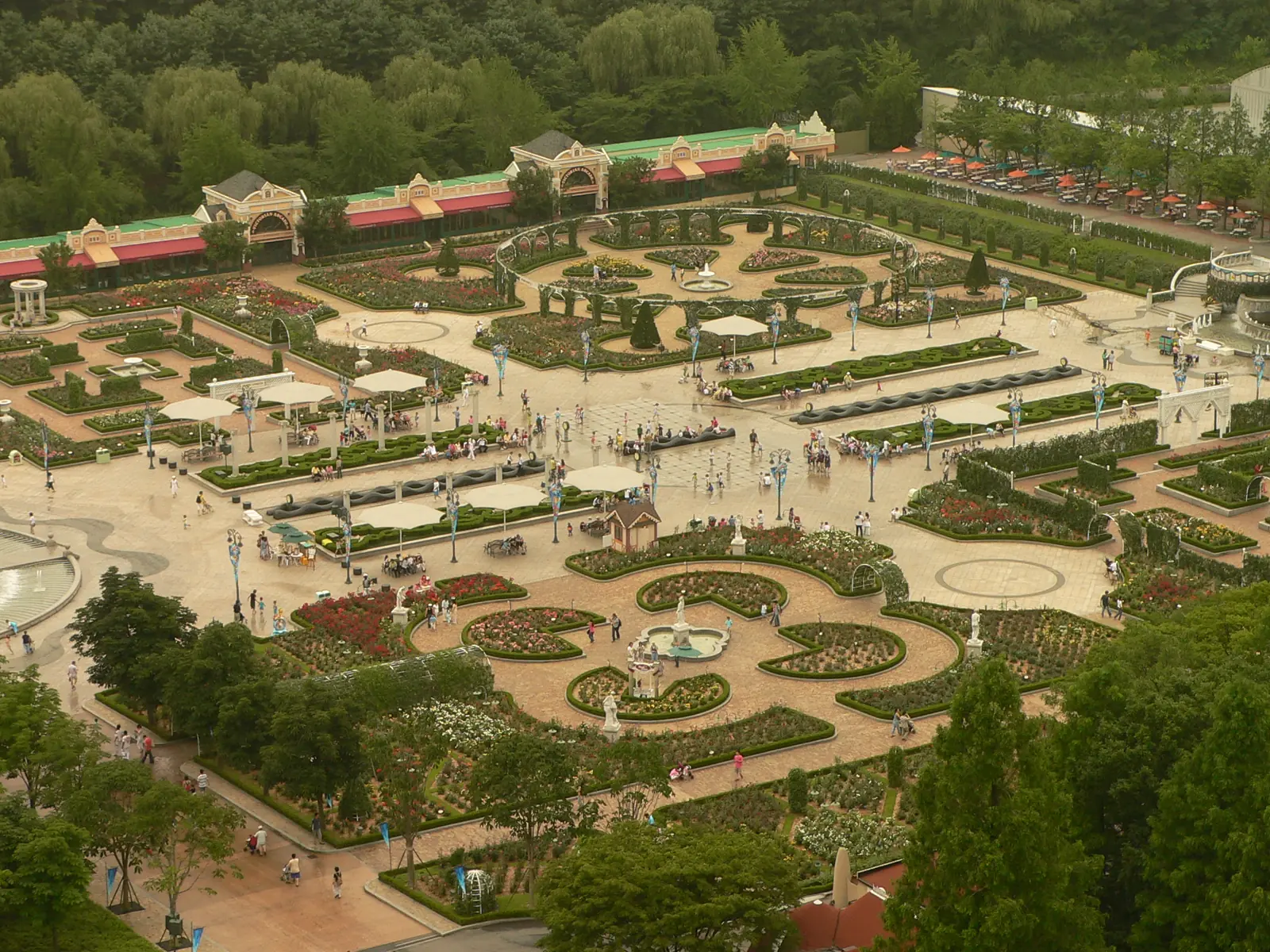 Jardines formales con senderos curvos, fuentes y sombrillas blancas vistos desde arriba, con un edificio largo y bajo y árboles al fondo