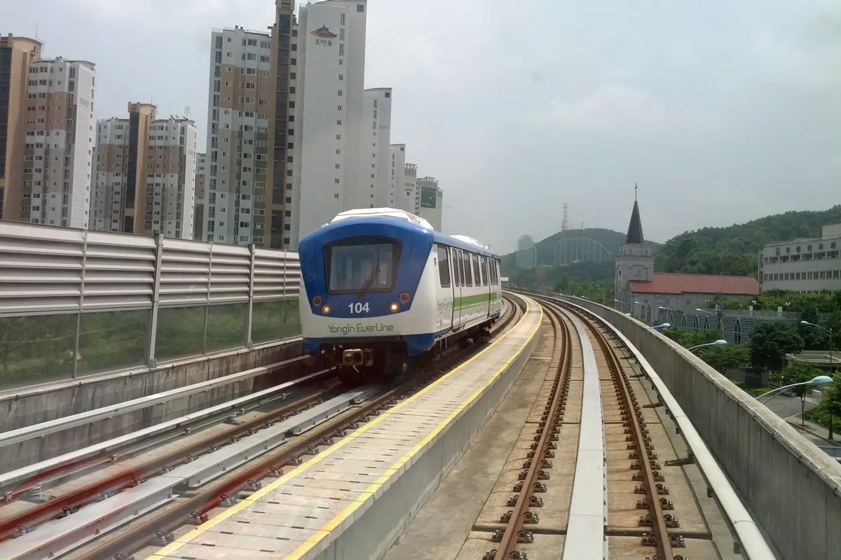 Un tren blanco y azul de Yongin Everline con el número 104 en su frontal circula por una vía elevada de hormigón junto a torres residenciales, con colinas boscosas al fondo