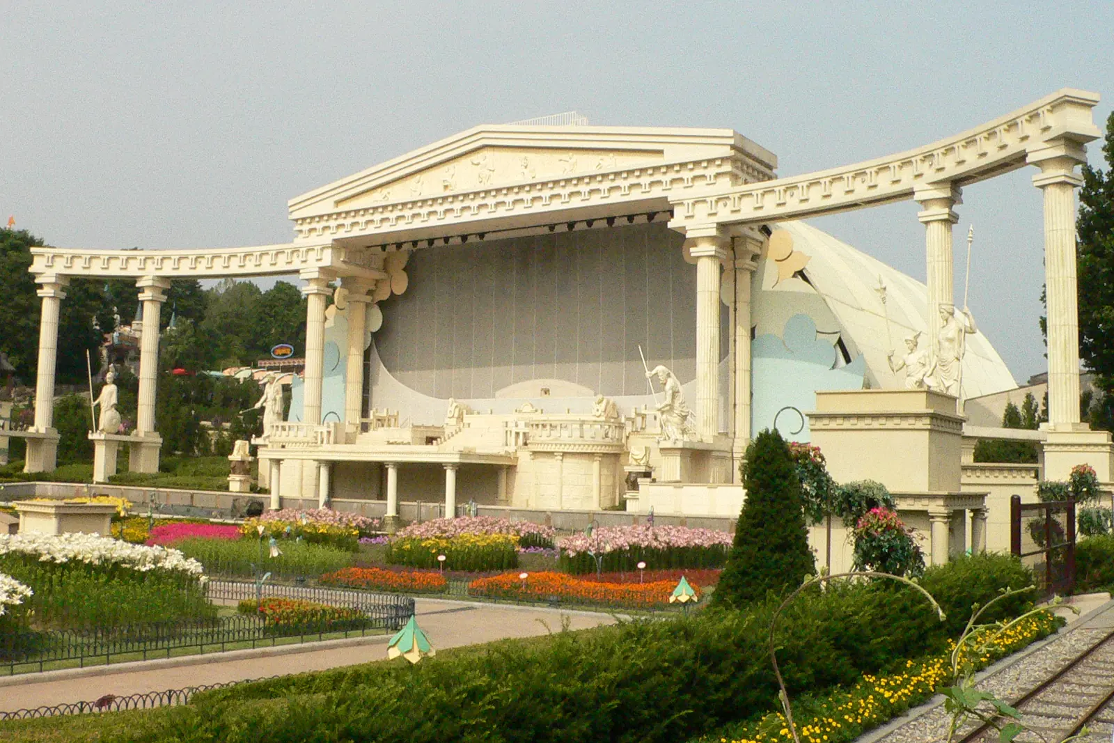 Un escenario blanco al aire libre con columnas y estatuas de estilo griego se alza tras parterres de flores y un seto recortado bajo un cielo brumoso